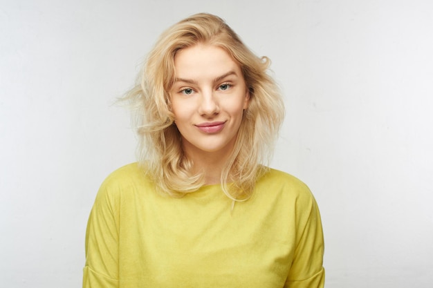 Studio shot van een jong aantrekkelijk blond meisje met onzorgvuldig golvend haar, een gele jas en een zachte glimlach op haar lippen, een portret op een witte achtergrond alleen