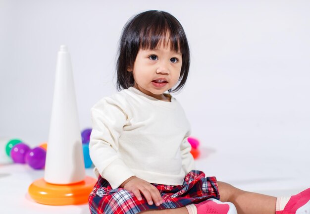 Studio shot van een klein schattig Aziatisch babymeisje dochtermodel in casual outfit zittend op de vloer terwijl ze spelen met kleurrijke plastic ringen constructie piramide speelgoed en kleine ballen op een witte achtergrond.