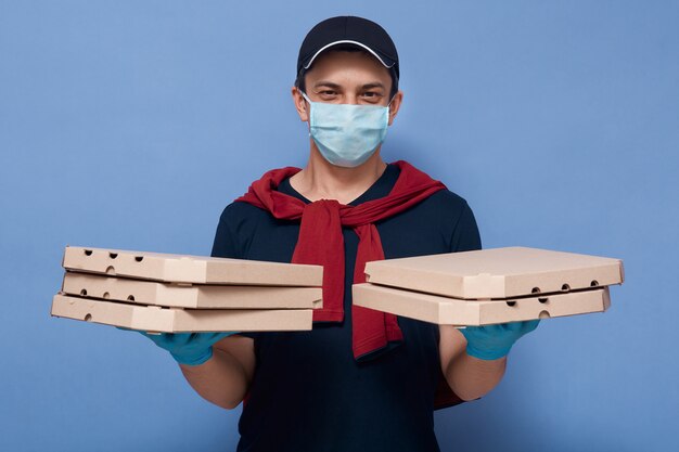 Studio shot van jonge knappe bezorger met kartonnen pizzadozen in beide handen, koerier jurken casual outfit en beschermend masker en handschoenen geïsoleerd over blauw