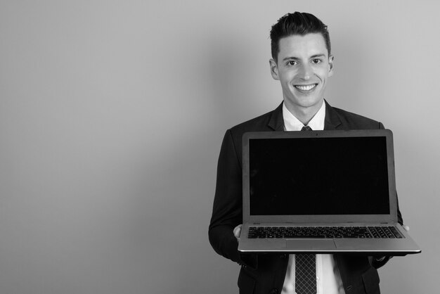 Studio shot van jonge knappe zakenman pak dragen terwijl laptop tegen grijze achtergrond in zwart-wit