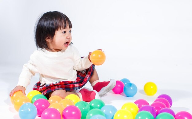 Studio shot van kleine schattige korte zwarte haren aziatische baby meisje dochter model in casual geruite rok zittend op de vloer lachend lachen spelen met kleurrijke ronde ballen speelgoed alleen op witte achtergrond.