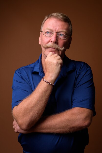 Studio shot van knappe senior man met snor dragen blauwe polo shirt en bril tegen bruin