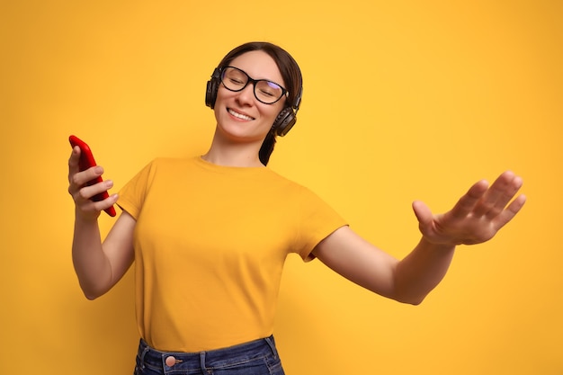 Studio shot van mooie brunette vrouw in geel t-shirt en draadloze koptelefoon luistert naar luide muziek en dansen op geel