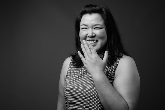 Studio shot van mooie dikke Aziatische vrouw, gekleed in jurk tegen een grijze achtergrond in zwart en wit