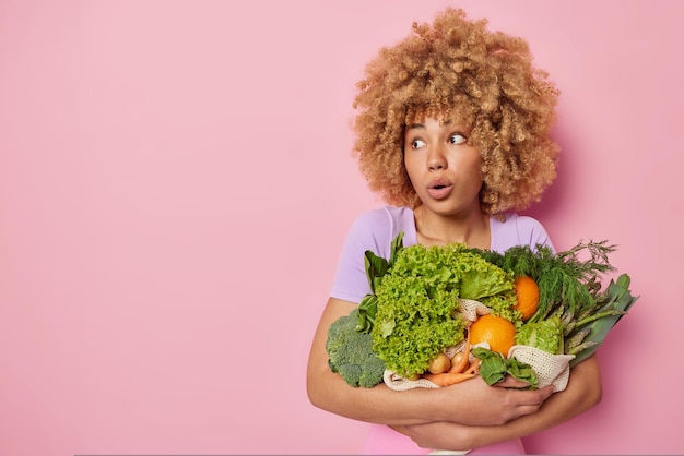 Foto studio shot van onder de indruk vrouw poseert met verse groene groenten geschokt om geweldig nieuws te horen gekleed in casual t-shirt geïsoleerd over roze achtergrond lege ruimte voor uw advertentie-inhoud