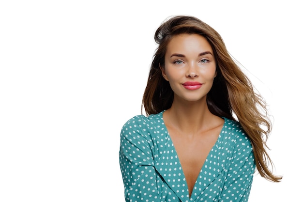 Studio shot van positieve jonge vrouw heeft lang golvend haar make-up draagt turquoise polkadot blouse kijkt recht naar cameramodellen tegen witte achtergrond kopieer ruimte voor uw advertentie