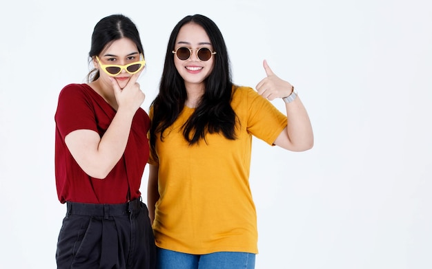 Studio shot van twee jonge Aziatische coole modieuze stijlvolle vrouwelijke vrienden in casual tshirt outfit dragen fashion vintage zonnebril staande glimlachend kijken elkaar ogen op witte achtergrond.