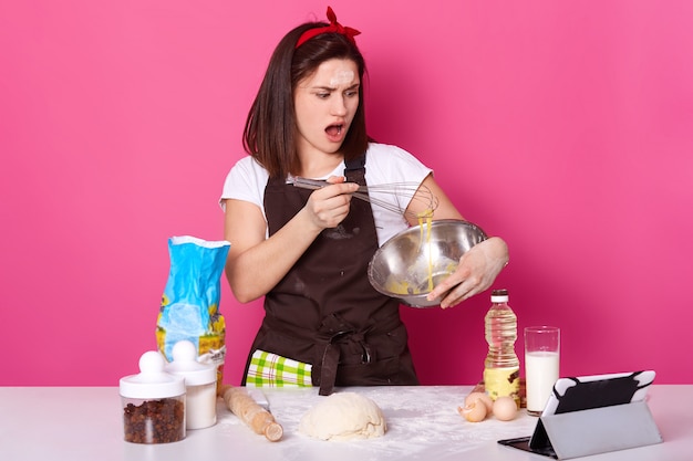 Studio shot of woman whisking eggs in kitchen, has astonished facial expression, making homemade pastry, baking cakes, stands near table, surrounded different producs. Baking and cooking concept.