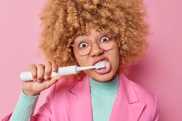 Studio shot of woman with curly hair cleans teeth uses electric toothbrush and fresh toothpaste does morning routine wears spectacles and elegant formal jacket Oral dental health toothcare concept