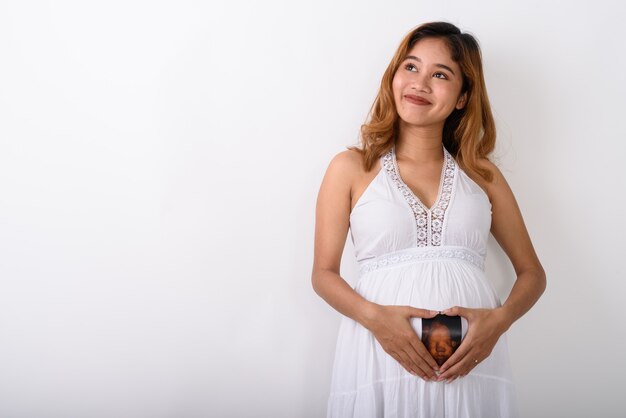 Studio shot of young Asian pregnant woman thinking while making