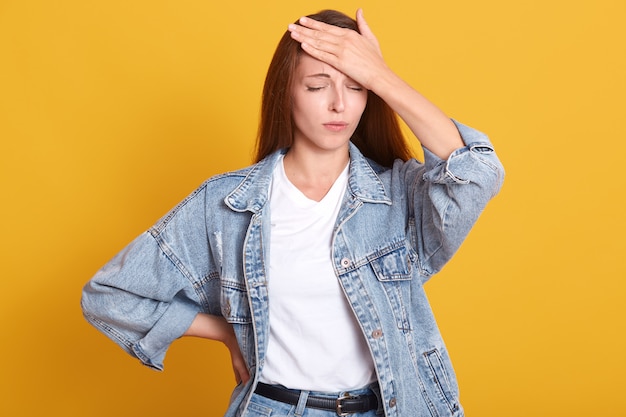 Lo studio ha sparato di giovane donna attraente isolata sopra il fondo giallo dello studio che sta con la mano sulla fronte, ricorda l'errore o qualcosa di importante.