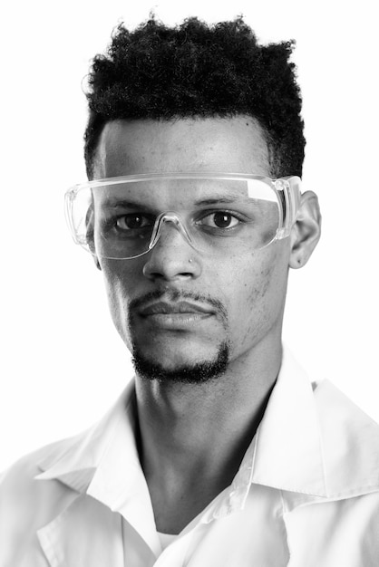 Studio shot of young bearded African man doctor with protective glasses isolated against white background in black and white