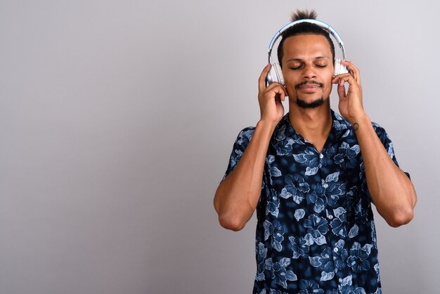 Studio shot of young bearded handsome African man wearing Hawaiian shirt while listening to music against gray background