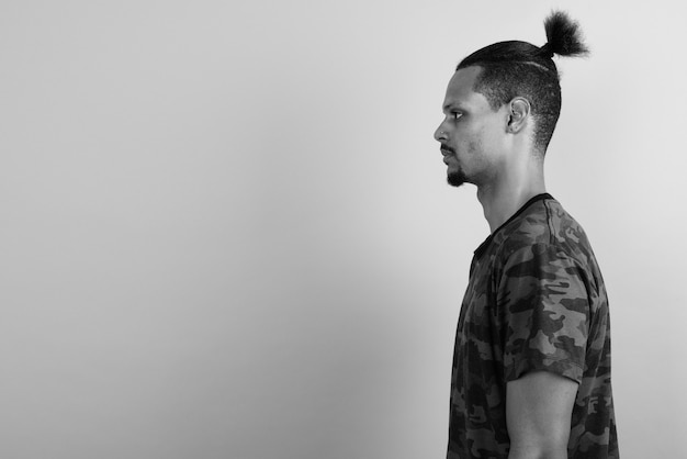 Studio shot of young bearded handsome African man with hair tied against gray wall in black and white