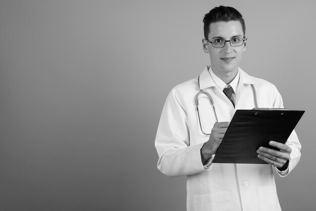 Studio shot of young handsome man doctor wearing eyeglasses against gray background in black and white