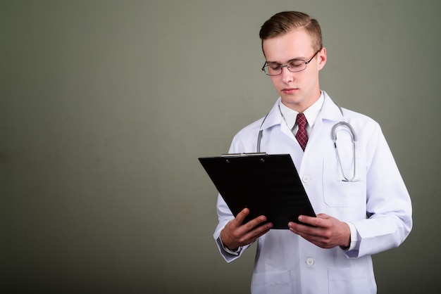 Studio shot of young handsome man doctor wearing eyeglasses while holding clipboard against colored background