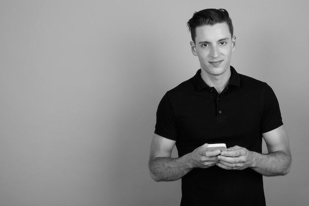 Studio shot of young handsome man using mobile phone against gray background in black and white