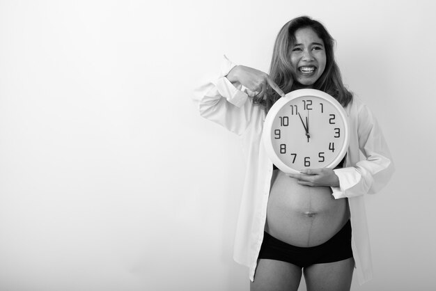 Studio shot of young happy Asian pregnant woman smiling while pointing at wall clock against white wall