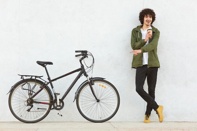 Studio shot of young male with curly hair, dressed in fashionable anorak, points with index finger at bicycle, advertises new model, drinks takeaway cooffe, makes choice, isolated on white background.