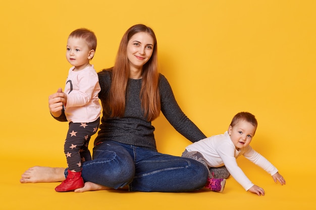 若い母親と彼女の双子の幼児のスタジオ撮影は、黄色で分離された写真スタジオでポーズします。ママは乳児と一緒に床に座り、大きな愛で抱きしめます。