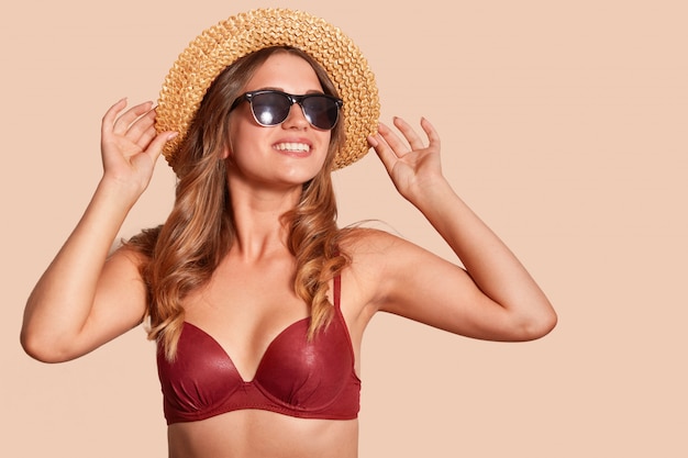 studio view of cheerful woman with toothy smile, wearing black sunglasses, straw hat, red swimming suit, looks in distance, isolated on beige, free space for your promotional content.