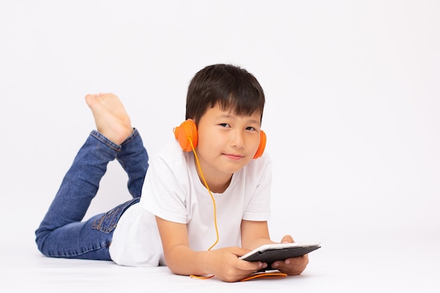 A studio view of a young preschool boy, laying on the floor and listening to music or a video on a tablet