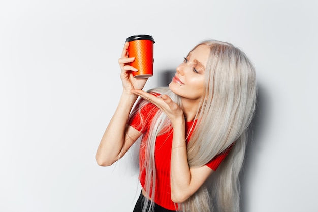 Studioportret van een jong glimlachend schattig meisje met blond haar met een papieren kopje koffie afhaalmaaltijden met een rood shirt op een witte achtergrond