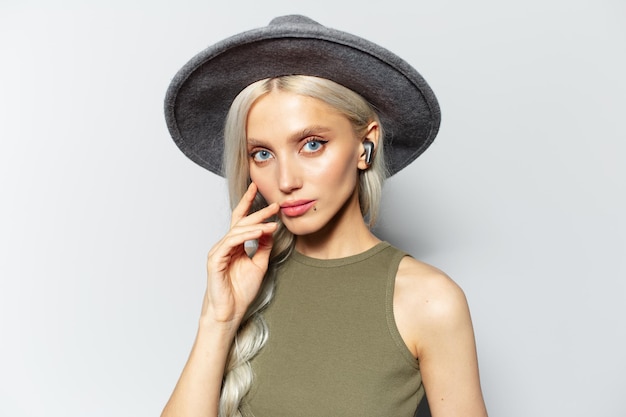 Studioportret van jong blonde aantrekkelijk meisje met oordopjes in oren op witte achtergrond die grijze hoed dragen
