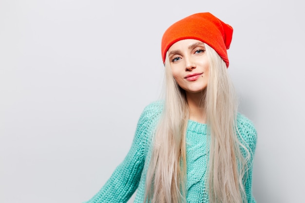 Foto studioportret van jong blondemeisje die oranje hoed en blauwe sweater op witte achtergrond dragen.