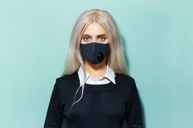 Studioportret van jong blondemeisje met blauwe ogen in schoolvorm, die zwart ademhalingsgezichtsmasker dragen tegen coronavirus op achtergrond van cyaan, aqua menthe-kleur.