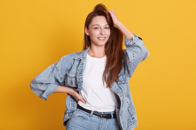 Studioportret van mooi vrouwenmodel met mooi donker lang haar, status dragend denimjasje en wit overhemd, geïsoleerd stellen over geel