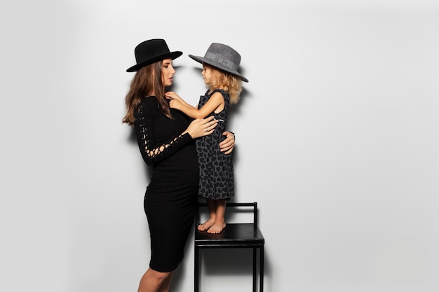 Studioportret van zwangere glimlachende vrouw en kindmeisje die elk ooit hoeden dragen op een witte achtergrond Moeder en dochter in het zwart