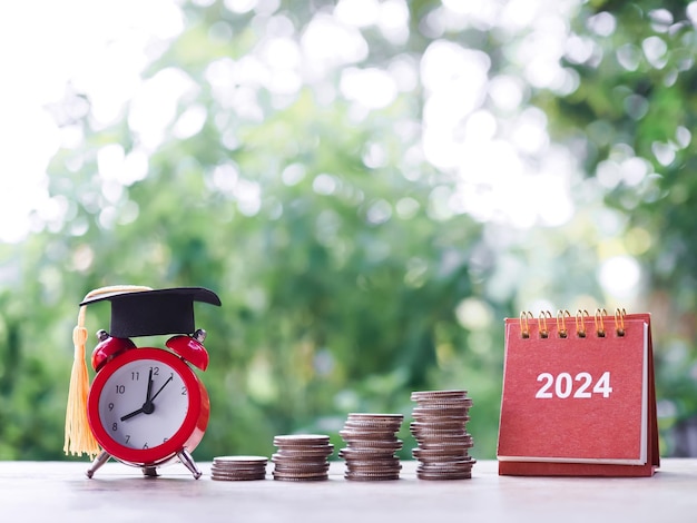 Фото Цели исследования 2024 настольный календарь красный будильник с выпускной шляпой и стопкой монет концепция экономии денег на образование студенческий кредит стипендия на оплату обучения в новом 2024 году
