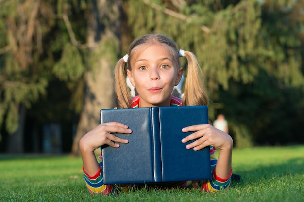 喜んで勉強してください。素晴らしい物語の時間。屋外で本を読んでいる小さな子供。女子高生は緑の芝生をリラックスしながら物語を読みます。かわいい生徒は読書を楽しんでいます。授業時間。子供のための興味深い話。