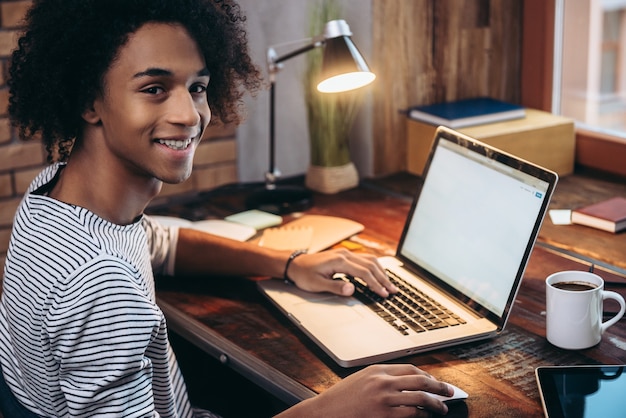 Studying at home. Side view of cheerful young African man working on laptop and looking at camera while sitting at his working place