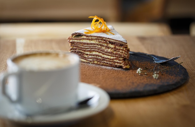 Stuk cacao gesneden cake op een leisteen ronde plaat met een kopje capuchino op een houten tafel
