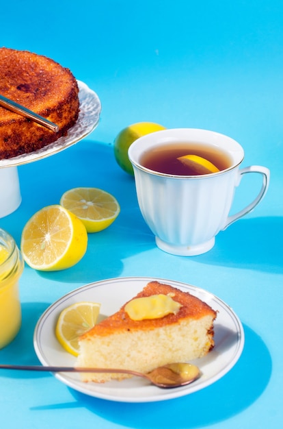 Stuk zelfgemaakte versgebakken biscuitgebak op plaat, Lemon Kurd in een glazen pot met lepel, hele en gesneden verse citroenen op plaat over blauwe achtergrond met schaduwen. Ruimte kopiëren. hard licht