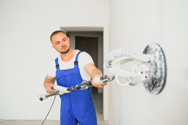 Stukadoor maakt het muuroppervlak glad met een muurschuurmachine. Meesterbouwer slijpt een witte gipsmuur. een man in overall maalt het oppervlak in een gasmasker. ervaren reparateur