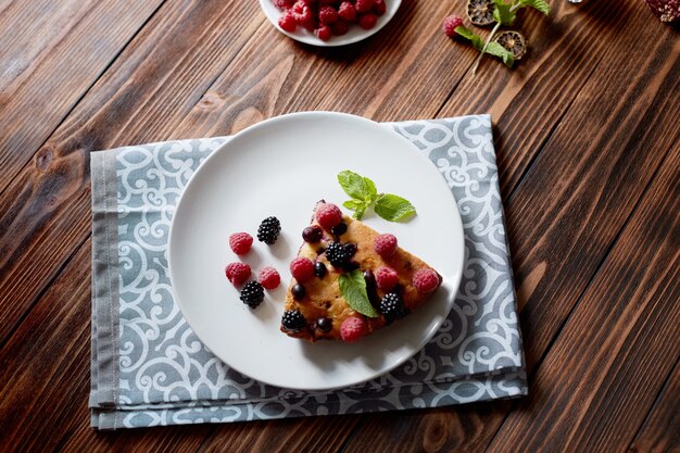 Stukje taart met bosbessen, frambozen en munt als dessert op een witte plaat, servet op een houten planken-tafel