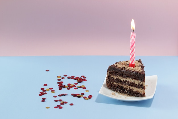 Stukje verjaardagstaart met een kaars en confetti op een blauwe achtergrond Stuk chocoladetaart