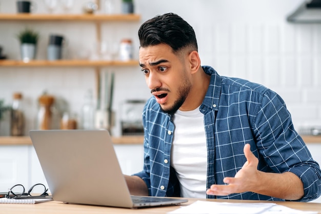 Stunned confused indian or arabian guy freelancer ort student sitting at home in the kitchen looking disappointment at the laptop screen stressed from the failure of a deal or project bad news