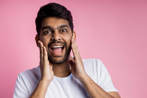 Stunned handsome unshaven indian male, being shocked by low prices on sale, keeping hands on cheeks, opening mouth in surprise, isolated over pink background. Emotions, reactions concept. Copy space.