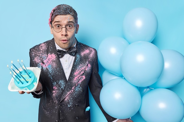 Stunned man celebrates birthday or other special occasion hlds festive sweet cake and bunch of inflated balloons gets unexpected congratulation isolated over blue background Festivity concept