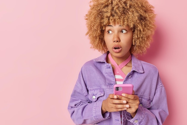 Stunned woman with curly hair receives email with bad news
feeling shocked holds mobile phone focused away dressed in purple
jacket isolated over pink background with copy space for your
advert