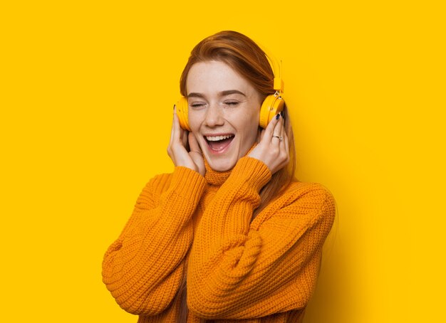 Stunning ginger caucasian lady is listening to music while wearing a sweater on a yellow background with copy space