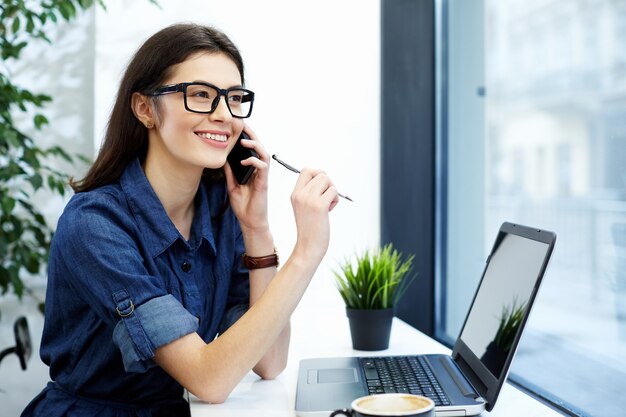 Stunning girl with black hair wearing shirt and eyeglasses sitting in cafe with laptop and cup of coffee, freelance concept, working process.