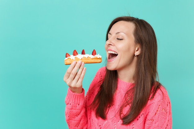 Foto splendida giovane donna gioiosa in maglione rosa lavorato a maglia che tiene in mano e mangia torta eclair isolata su sfondo blu turchese parete, ritratto in studio. concetto di stile di vita della gente. mock up copia spazio.