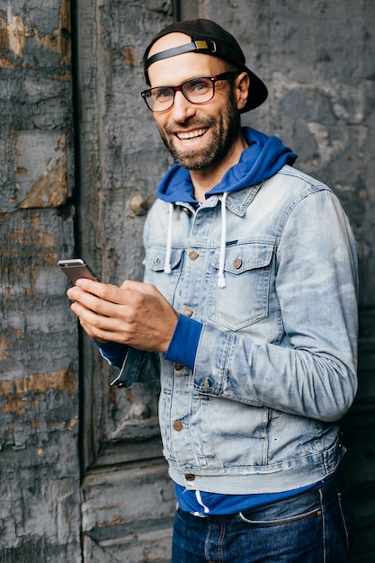 Stunning man with blue eyes, beard and broad smile wearing cap, denim shirt and eyeglasses holding smartphone