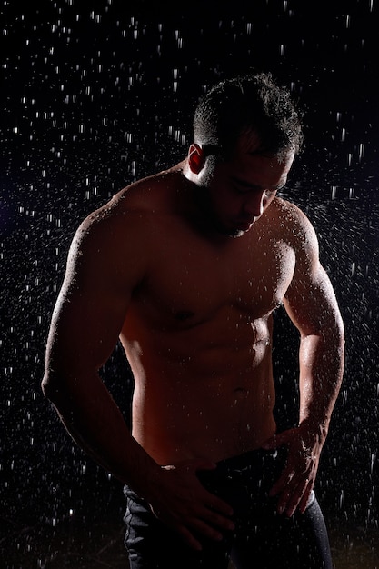 Stunning man with muscular body posing in rain, with naked torso, water drops on body