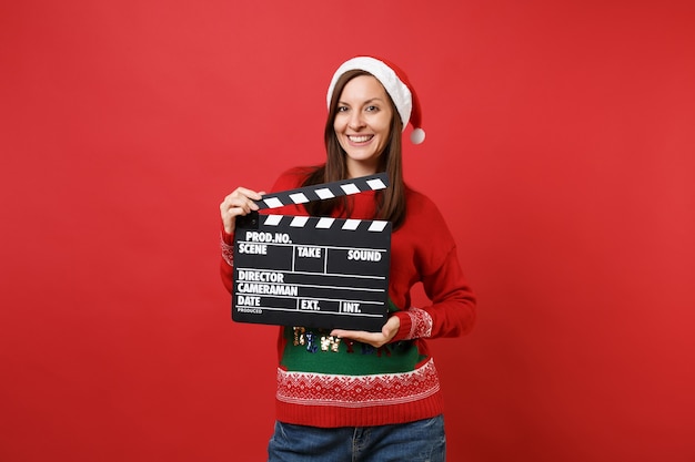 Stunning merry young Santa girl in Christmas hat holding classic black film making clapperboard isolated on red background. Happy New Year 2019 celebration holiday party concept. Mock up copy space.